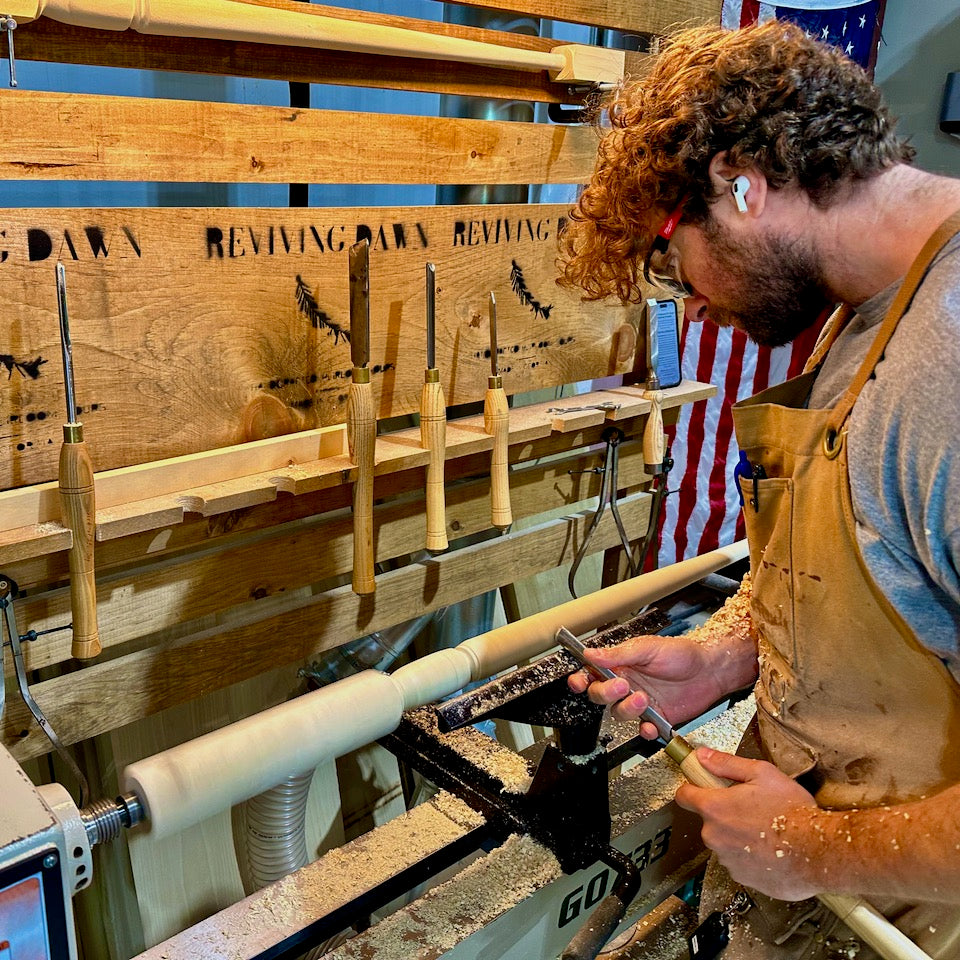 Kyle Oliver turning a pintop baluster at the lathe, the Reviving Dawn tool rack behind him